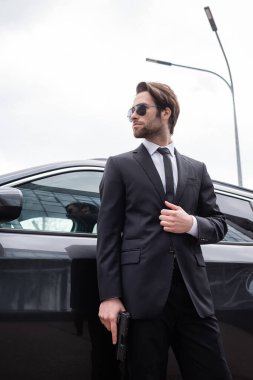 low angle view of bearded safeguard in sunglasses and suit standing near modern car