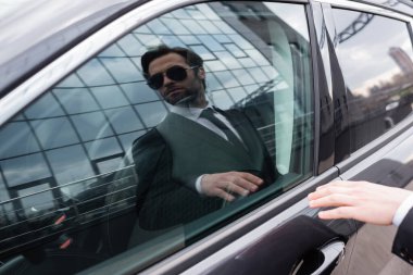 reflection of bearded safeguard in sunglasses and suit opening door in modern car