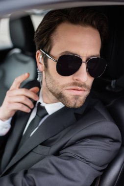 bearded bodyguard in sunglasses and suit adjusting security earpiece in car