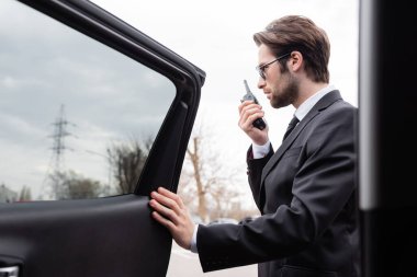 side view of bearded bodyguard in suit and sunglasses using walkie talkie near modern car