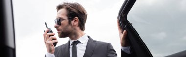 side view of bearded bodyguard in suit and sunglasses using walkie talkie near modern car, banner