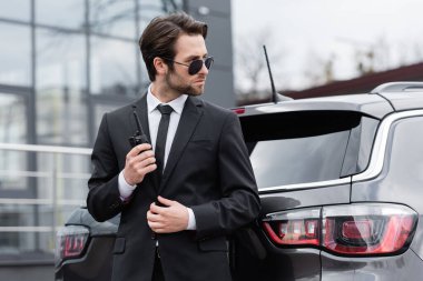 side view of bodyguard in sunglasses holding walkie talkie near modern car