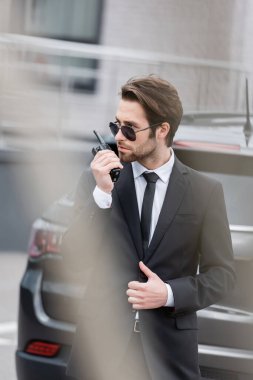 bodyguard in sunglasses and suit using walkie talkie near blurred modern car