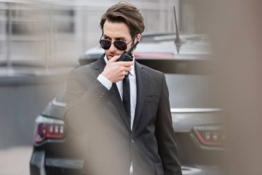 bodyguard in sunglasses and suit using modern walkie talkie near blurred car