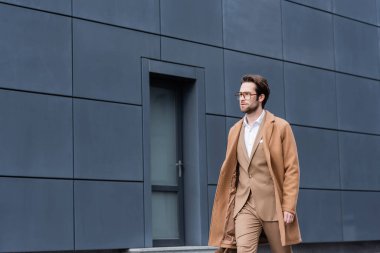 bearded businessman in glasses and beige coat walking near building outside