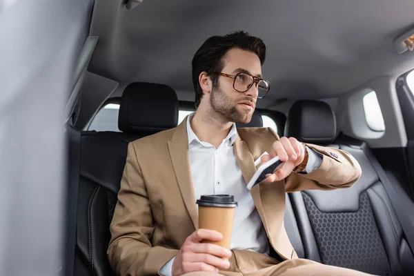 身穿米黄色西服 戴着眼镜的商人在车里拿着纸杯和智能手机 — 图库照片