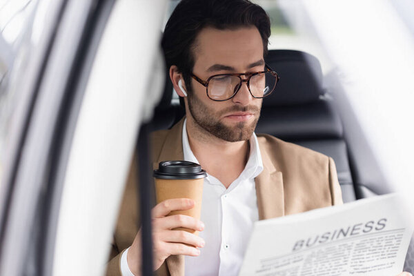 bearded man in suit reading business newspaper and holding coffee to go in car 