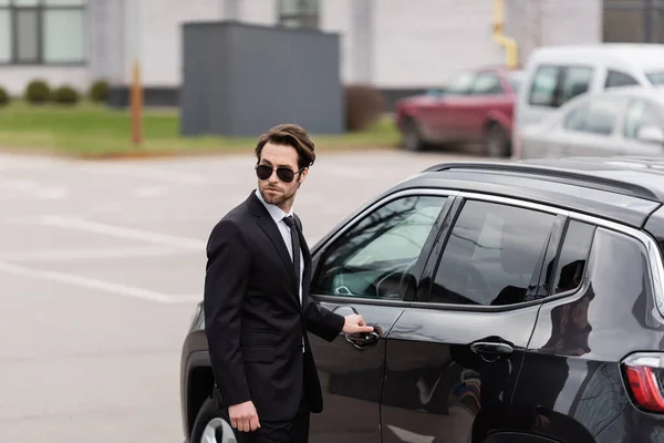 bodyguard in suit and sunglasses with security earpiece opening door of modern auto