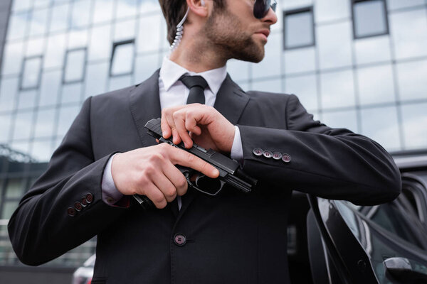 side view of bearded bodyguard in suit holding gun near modern building 