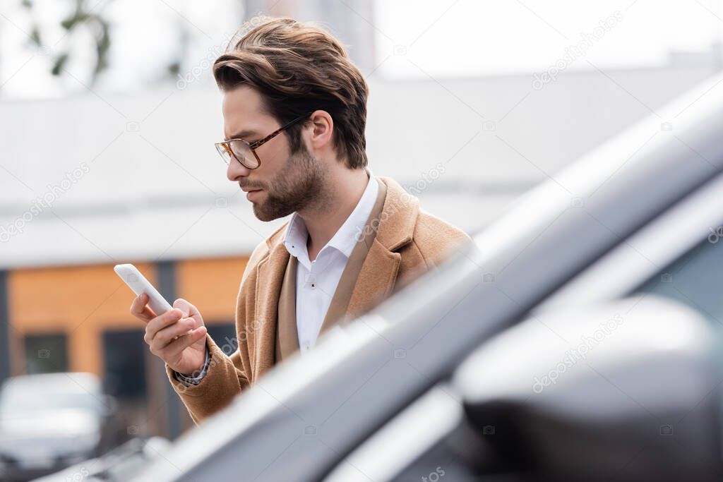 young man in glasses and beige coat looking at cellphone near blurred car