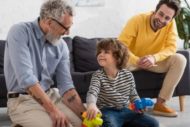 Mature man looking at grandson with colorful building blocks near son at home  clipart
