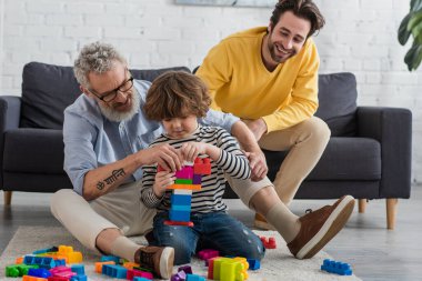 Kid stacking building blocks near grandfather and father in living room  clipart