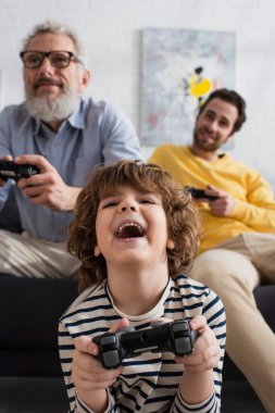 KYIV, UKRAINE - APRIL 12, 2021: Excited kid playing video game near blurred parents  clipart