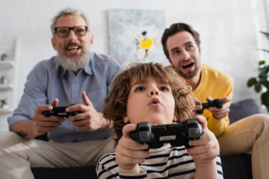 KYIV, UKRAINE - APRIL 12, 2021: Low angle view of excited child playing video game near father and granddad on blurred background  clipart