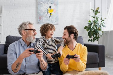 KYIV, UKRAINE - 12 Nisan 2021: Neşeli çocuk dedesinin yanında babasına joystick ile bakıyor 