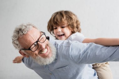 Cheerful granddad playing with blurred grandson at home clipart