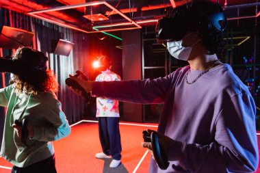 Tıp maskeli çok ırklı genç arkadaşlar kulaklıkla VR oyunu oynuyorlar.
