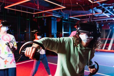 VR kulaklıklı ve tıbbi maske takan Afrikalı Amerikalı kız, bulanık arkadaşlarının yanında oyun oynuyor.