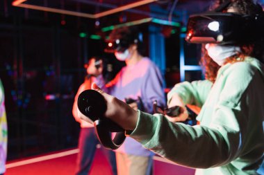 african american teenage girl in vr headset and safety mask gaming near blurred friends clipart
