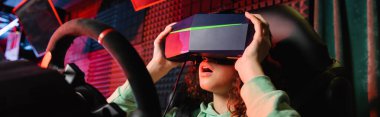 Araba simülatörü ve pankartta oynarken VR kulaklığı ayarlayan Afrikalı Amerikalı kız hayrete düştü.