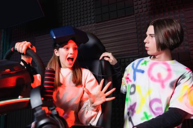 amazed girl on car simulator gesturing near surprised friend clipart