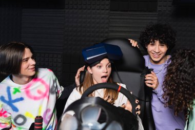 multiethnic friends laughing near astonished girl on car racing simulator clipart
