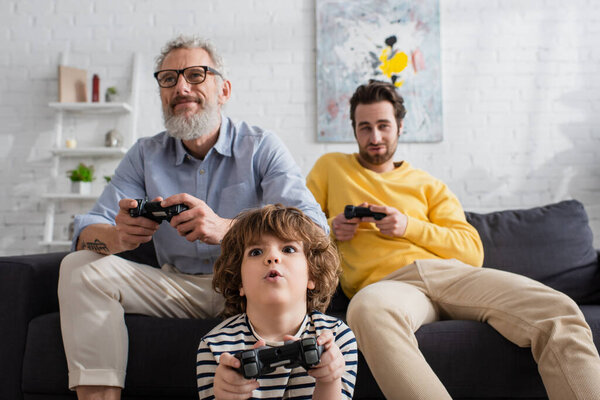 КИЕВ, УКРАИНА - 12 апреля 2021 года: Удивительный мальчик играет в видеоигры возле размытых мужчин на диване 