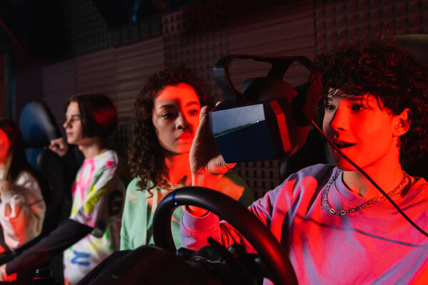 amazed teenager on car simulator holding vr headset near african american friend