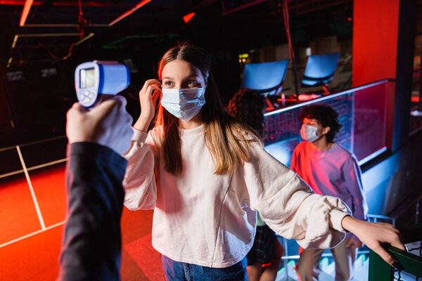 teenage girl in medical mask near controller with pyrometer and friends on blurred background