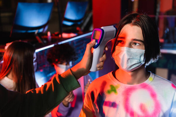 gamer in medical mask near girl with pyrometer and blurred friends on background