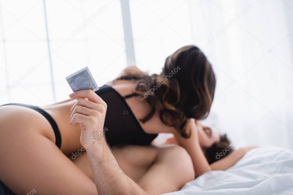 Condom in hand of man kissing sexy girlfriend on blurred background in bedroom 