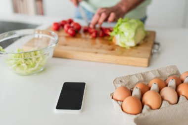 Tavuklu yumurta ve akıllı telefonun seçici odağı. Bulanık adamın yanında kahvaltı hazırlıyor. Kesilmiş manzara.