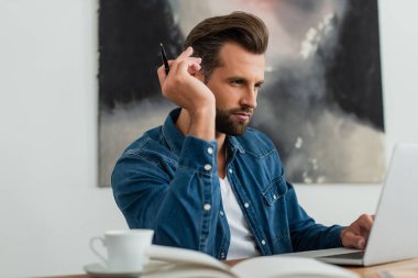 Önplanda bulanık dizüstü bilgisayarın yanında düşünürken elinde kalem tutan yetişkin bir adam