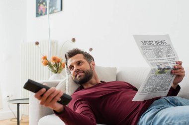 Kanepede gazeteyle yatarken televizyon kanallarını tıklatan neşeli bir adam.