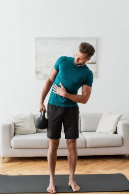 Çıplak ayaklı adamın oturma odasında kettlebell kaldırışının geniş açılı görüntüsü.