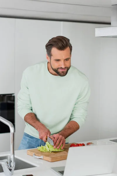 Homme Souriant Regardant Ordinateur Portable Tout Coupant Des Légumes Dans — Photo