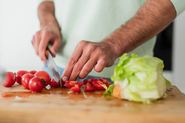cropped view of man cutting cherry tomatoes near fresh lettuce in kitchen
