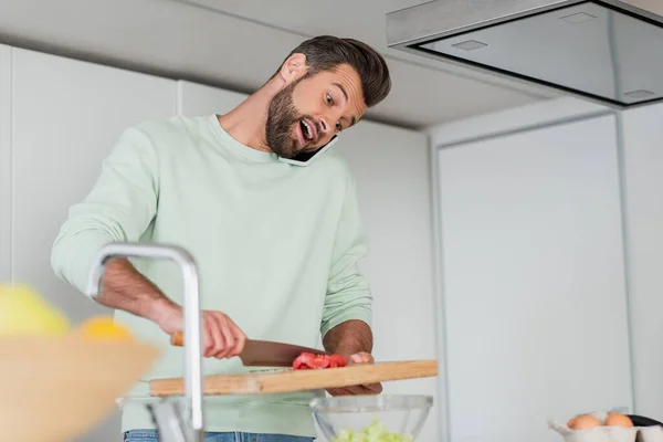 amazed man talking on smartphone while preparing salad on blurred foreground