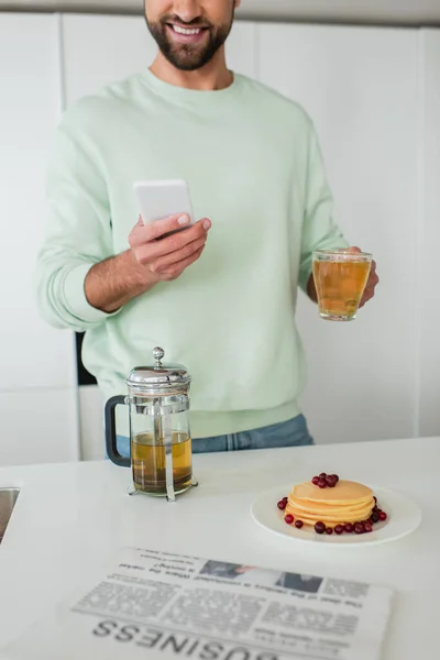 キッチンでパンケーキの近くでスマホと緑茶を持っている笑顔の男の部分的な眺め — ストック写真