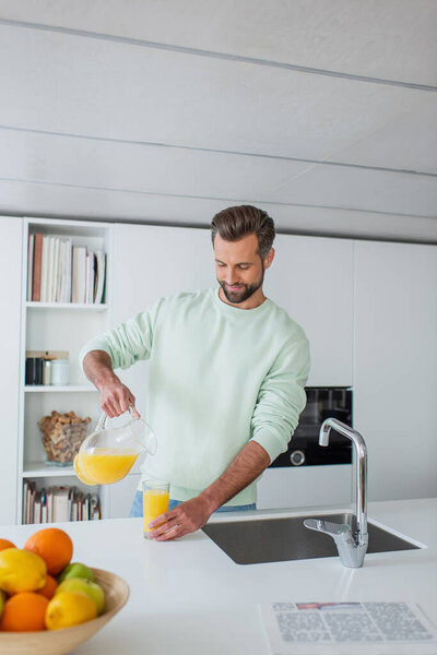 positive man pouring orange juice near fresh fruits on blurred foreground in kitchen