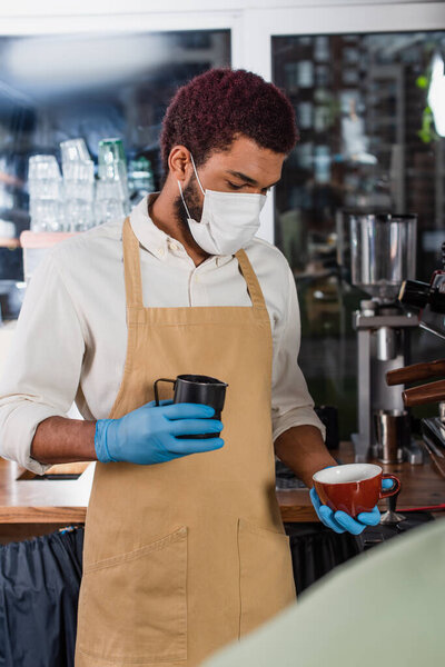 Африканская американская бариста в медицинской маске держит чашку и молочный кувшин рядом с кофеваркой 