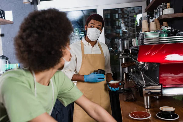 African american barista in medical mask and latex gloves making coffee near blurred colleague in cafe
