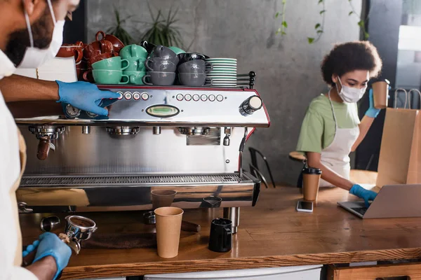 African american barista in medical mask and latex gloves using coffee machine near blurred colleague with laptop