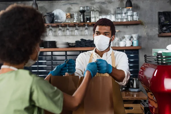 Tıbbi Maskeli Afrikalı Amerikalı Barista Bulanık Önplandaki Meslektaşına Kağıt Torba — Stok fotoğraf