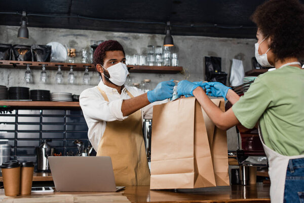 Африканский американец-бариста в медицинской маске дает бумажные пакеты коллеге возле кофе, чтобы пойти и ноутбук 