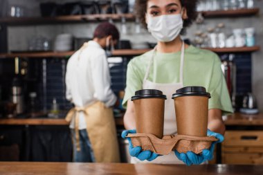 Kağıt bardaklar bulanık arka planda tıbbi maske takan Afrikalı Amerikalı baristanın ellerinde. 