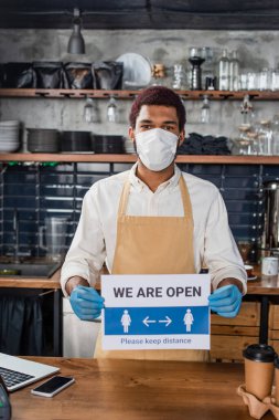 Tıbbi maskeli Afrikalı Amerikalı barista ve levha tutan lateks eldivenli kahve dükkanında açık harf kullanıyoruz. 