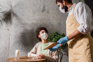 Tıbbi maskeli Afrikalı Amerikalı garson müşterinin yanında menüyü tutuyor. 