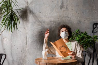 Kafe 'de akıllı telefonun yanında tıbbi maskeli Afrikalı Amerikalı bir kadın menüyü tutuyor. 