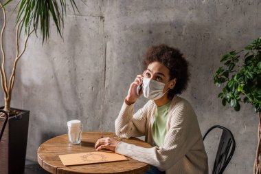 Tıbbi maskeli Afrikalı Amerikalı bir kadın kafenin yakınlarında akıllı telefondan konuşuyor. 